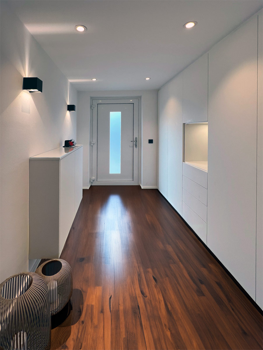 Dieser Flur zeigt ein Zusammenspiel aus Stauraum und modernem Lichtdesign. Auf der rechten Seite fügt sich ein deckenhoher Einbauschrank in Weiß nahtlos in die Architektur ein. Eine beleuchtete Nische unterbricht die geschlossene Front und dient als praktische Ablage. Gegenüber bietet ein halbhohes Sideboard zusätzlichen Platz, während dunkle Holzdielen und schwarze Wandleuchten einen eleganten Kontrast zu den hellen Oberflächen bilden.